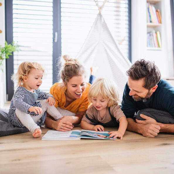 Junge Familie mit zwei kleinen Kindern sitzt drinnen auf dem Boden liest gemeinsam ein Buch.